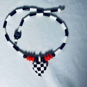 May include: A black and white checkered heart pendant necklace with red beads and a black and white bead chain. The pendant has the word "Loved" written on it.