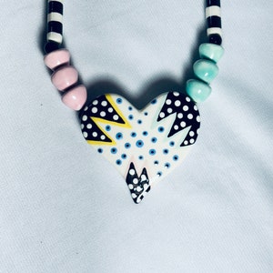 May include: A black and white striped necklace with pink and blue beads and a heart-shaped pendant. The pendant is white with black polka dots, yellow accents, and blue and white swirls.