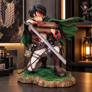 Op de afbeelding: Een gedetailleerd anime-figuur met zwart haar, gekleed in een bruin jasje, witte broek en een groene cape, knielend op een gestructureerde basis. Het figuur houdt een zilveren zwaard vast en is bespat met rode verf die op bloed lijkt.