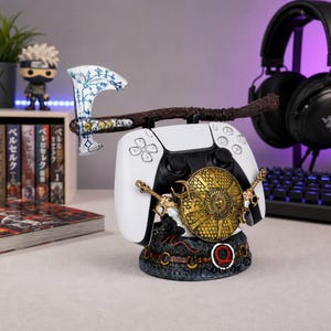 May include: A white and black video game controller stand featuring a decorative axe with blue markings, a gold circular shield, and a dark base with a red Omega symbol. The stand is designed to hold a controller securely while displaying themed elements.