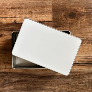 May include: A white rectangular box with a silver trim sits open on a wooden surface. The box is empty.