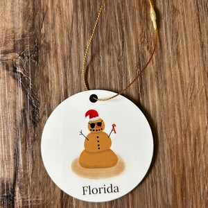 May include: Round white ornament with a brown snowman wearing sunglasses and a red Santa hat. The snowman is standing in the sand with a red candy cane. The ornament says "Florida" at the bottom.