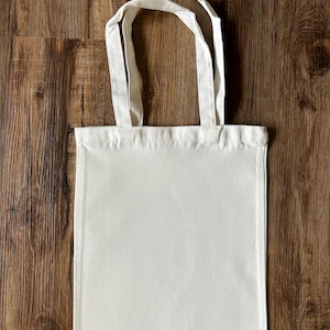 May include: A plain, cream-coloured tote bag with two matching handles. The bag has a rectangular shape and a simple design, suitable for carrying items. The background is a wooden surface.