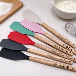 May include: A set of five silicone spatulas with wooden handles, each featuring unique laser-engraved text including Kendia's Kitchen, With Love, Have a nice day, Made with love & Kendia, Grandma's Kitchen, and Teneisha with a heart symbol.