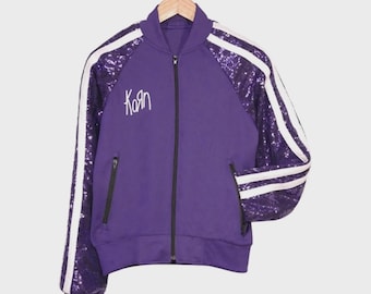 Könnte beinhalten: Eine violette Bomberjacke mit Reißverschluss, langen Ärmeln mit violetten Pailletten und zwei weißen Streifen an jedem Arm. Auf der Vorderseite befindet sich der weiße Schriftzug Korn, ein schwarzer Reißverschluss und zwei Reißverschlusstaschen.