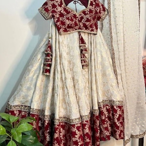 以下が含まれることがあります： 深いマルーン色のブラウスに金色の花柄刺繍が施され、幅広のマルーン色の縁取りがある白いスカートとセットになったツーピース。スカートとブラウスには複雑な金色の模様があり、金色のドットが入った透け感のある白いデュパタが含まれています。