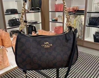 May include: A dark brown Coach shoulder bag featuring the signature monogram pattern, a gold-tone zip closure, and a gold-tone chain link handle. The bag includes a detachable black leather strap and a gold-tone Coach logo on the front.
