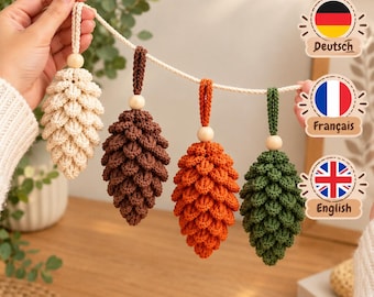 May include: A garland featuring four crocheted pinecone ornaments in cream, brown, orange, and green. Each pinecone is suspended from a cream-coloured rope by a small wooden bead and a matching crocheted loop, creating a textured, seasonal display.