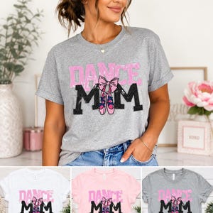 May include: A heather grey short-sleeve t-shirt featuring the text DANCE MOM in a sequined style. The word DANCE is in pink, and MOM is in black. A pink bow and a pair of pink ballet slippers are positioned between the letters M and M.