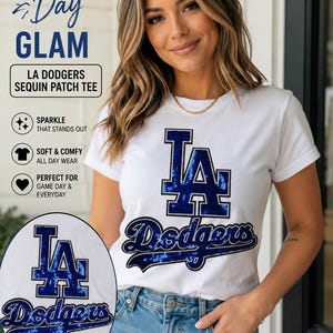 May include: A white short-sleeved t-shirt featuring a blue sequin LA Dodgers logo patch on the chest. The text on the shirt reads LA Dodgers. The design includes a large block letter LA above the script word Dodgers, both filled with blue sequins.