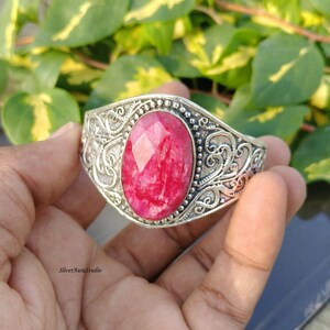May include: A silver cuff bracelet featuring an oval, faceted, vibrant pink gemstone at the centre. The metal band is decorated with intricate, raised scrollwork patterns and a beaded border surrounding the central stone. The text SliverAuraStudio is visible.