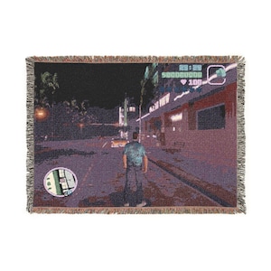 May include: A rectangular woven throw blanket featuring a pixelated video game scene at night. A character in a blue shirt and jeans stands on a street near a building with a neon sign. The display includes a circular map, health bar, and text.