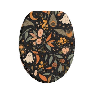 Op de afbeelding: Een ovale toiletbrilhoes met een zwarte achtergrond en een herhalend patroon van witte geesten, bruine vleermuizen, oranje maansikkels, gele sterren en diverse botanische elementen zoals bladeren en bloemen in gedempte tinten.