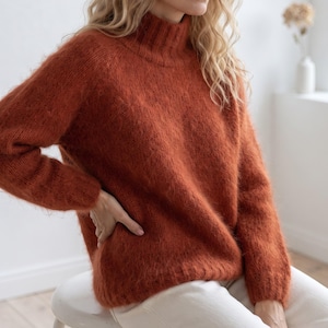 May include: A rust-colored, long-sleeved sweater with a high mock neck collar. The garment features a fuzzy, textured knit construction with raglan sleeves and ribbed detailing at the collar, cuffs, and hem, providing a soft, voluminous appearance.