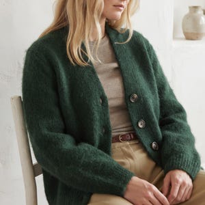 May include: A dark forest green long-sleeve cardigan with a fuzzy, textured knit finish. The garment features a V-neckline and is fastened with four large, round, dark-toned buttons down the front. It is worn over a beige ribbed top and tan trousers.