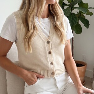 May include: A cream-colored knit sweater vest featuring a V-neckline, a button-down front with four brown buttons, and ribbed trim along the armholes, neckline, and hem. The vest is worn over a plain white short-sleeved t-shirt.