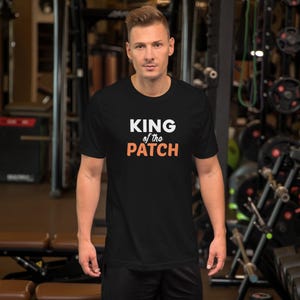 May include: A black short-sleeved t-shirt featuring the text KING of the PATCH printed on the front. The word KING is in large white capital letters, the word of the is in smaller white lowercase letters, and the word PATCH is in large orange letters.