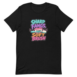 May include: A black short-sleeve t-shirt featuring a graphic of pink lips with sharp white fangs holding a light blue toothbrush. The text reads SHARP FANGS, in light blue and purple, and SOFT BRUSH, in orange and pink, centered on the chest.