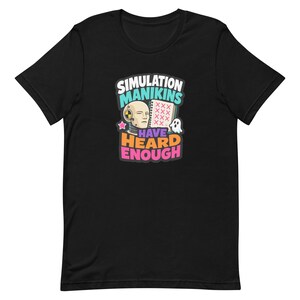 Puede incluir: Una camiseta negra de manga corta con un estampado gráfico de una cabeza de maniquí de pruebas de choque, un cuaderno de espiral con marcas de X rojas y un pequeño fantasma. El texto dice SIMULATION MANIKINS HAVE HEARD ENOUGH en letras blancas, turquesas, naranjas y rosas.