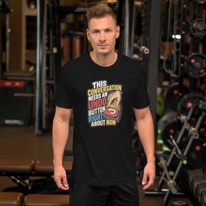 Op de afbeelding: Een zwart t-shirt met korte mouwen en een grafische print van een hand die op een rode knop drukt. De tekst luidt: THIS CONVERSATION NEEDS AN UNDO BUTTON RIGHT ABOUT NOW. Het ontwerp gebruikt gele, rode en blauwe letters op een donkere achtergrond.