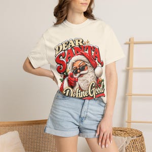 May include: A cream-colored short-sleeve t-shirt featuring a graphic of Santa Claus wearing sunglasses and pointing forward. The text reads Dear Santa, Define Good in red and black lettering with snowy accents and holly leaves.