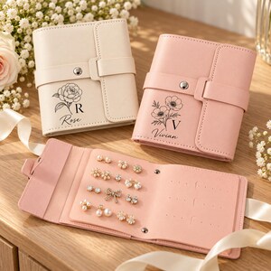 May include: Two rectangular jewelry organizers, one cream and one pink, each featuring a snap-button strap and floral line art with names Rose and Vivian. An open pink organizer displays various stud earrings and pearl-style jewelry pieces.