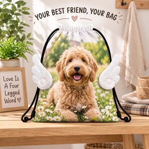 May include: A drawstring bag featuring a golden-brown dog lying in a field of white daisies. The bag has white plush angel wings attached to the sides and black drawstrings. The text "YOUR BEST FRIEND, YOUR BAG" is printed at the top.