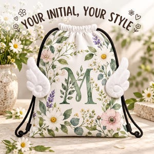 May include: A white drawstring bag featuring a large green letter M, decorated with watercolor-style flowers and foliage. The bag has two white plush angel wings attached to the sides and black drawstrings. Text reads: YOUR INITIAL, YOUR STYLE.