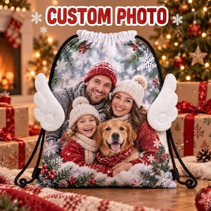 May include: A white drawstring backpack featuring a custom photo of a family and a golden retriever wearing winter clothing. The bag has white plush angel wings on the sides and a festive border of pine branches, red berries, and snowflakes.