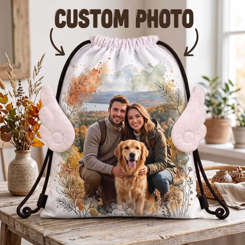 May include: A white drawstring backpack featuring a custom photo of a man, a woman, and a golden retriever dog. The bag is decorated with watercolor-style floral accents and two plush white angel wings attached to the sides. The text CUSTOM PHOTO is printed above the bag with arrows pointing down.