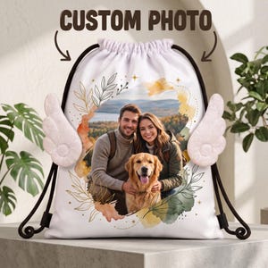 May include: A white drawstring backpack featuring a custom photo of a man, a woman, and a golden retriever. The bag has black drawstrings and two plush white angel wings attached to the sides. The photo is framed by watercolor floral art.