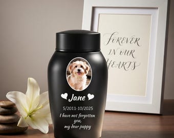 May include: A matte black pet cremation urn featuring a circular photo of a light-colored puppy, the name Jane, the dates 5/2011-10/2025, and the text I have not forgotten you, my dear puppy, accented by two white heart icons.