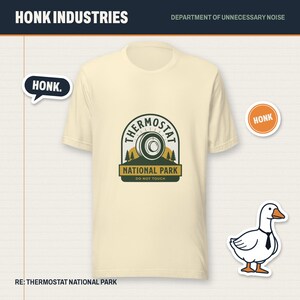 May include: A cream-colored short-sleeve t-shirt featuring a graphic of a thermostat dial above the text THERMOSTAT NATIONAL PARK DO NOT TOUCH. The design includes pine trees and a sun. A white goose wearing a black tie is also visible.