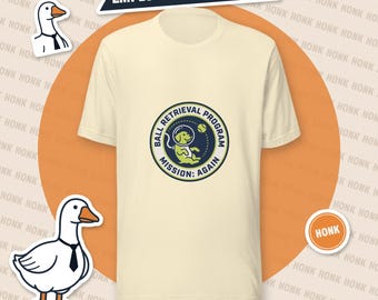 May include: A cream-colored t-shirt featuring a circular graphic with a dog in a space helmet, a tennis ball, and the text "BALL RETRIEVAL PROGRAM MISSION: AGAIN". The shirt is surrounded by two goose illustrations wearing ties and the text "EMPLOYEE OF THE MONTH" and "HONK".