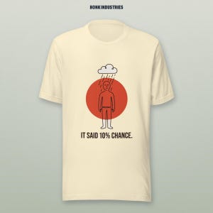 May include: A cream-colored short-sleeve t-shirt featuring a graphic of a person standing under a rain cloud in front of a red circle. The text "HONK INDUSTRIES" appears at the top, and "IT SAID 10% CHANCE." is printed below the graphic.