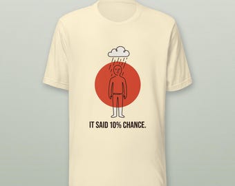May include: A cream-colored short-sleeve t-shirt featuring a graphic of a person standing under a rain cloud in front of a red circle. The text "HONK INDUSTRIES" appears at the top, and "IT SAID 10% CHANCE." is printed below the graphic.