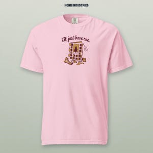 May include: A light pink short-sleeve t-shirt featuring a graphic of an open advent calendar with the text 'i'll just have one.' and 'DEC 1' on a small tag. The design includes a central Christmas tree illustration and scattered paper scraps.