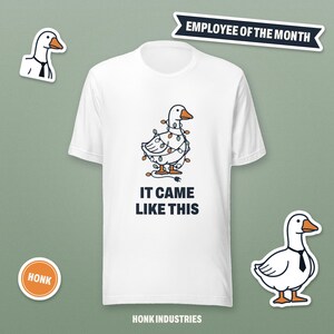 May include: A white short-sleeve t-shirt featuring a graphic of a goose wrapped in colorful string lights. Below the goose, the text reads IT CAME LIKE THIS. Additional elements include a goose with a tie, a HONK badge, and EMPLOYEE OF THE MONTH text.