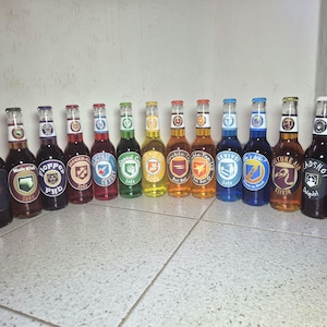 Könnte beinhalten: Eine Sammlung von vierzehn Glasflaschen gefüllt mit verschiedenfarbigen Flüssigkeiten, jede mit einem einzigartigen runden Etikett mit Aufschriften wie Mule Kick, Flopper PHD, Juggernog, Electric Cherry, Speed Cola, Double Tap, Revive und Tombstone Soda.
