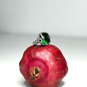 May include: A silver ring featuring a large, faceted emerald green gemstone set within a band designed to resemble a coiled snake with textured scales, resting atop a whole, round, deep red pomegranate fruit with a textured, leathery skin.