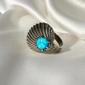 Pode incluir: Um anel de tom prateado com um design de concha texturizada em forma de leque. O centro do anel apresenta uma pedra cabochão redonda de um azul vibrante, exibindo reflexos iridescentes de verde brilhante e azul na sua superfície polida.