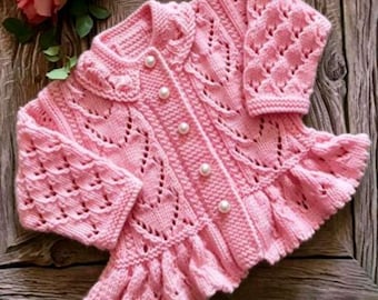 May include: A light pink long-sleeve knit cardigan featuring a delicate openwork leaf pattern, a ruffled hem, and five round white pearl-like buttons down the center front. The garment has a rounded collar and textured knit sleeves.