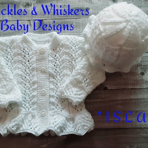 May include: White crocheted baby cardigan and matching hat set. The cardigan has a button fastening and a frilled hem. The hat is a simple, round shape. The set is labelled "Isela".