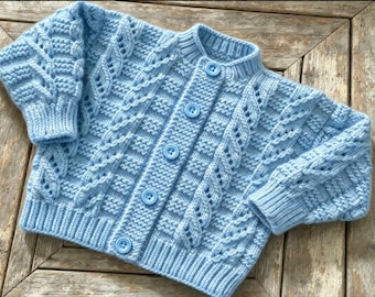 May include: A light blue knitted cardigan featuring a textured cable knit pattern, a ribbed collar, ribbed cuffs, and a ribbed hem. The front of the garment is secured with five matching light blue circular buttons aligned vertically.