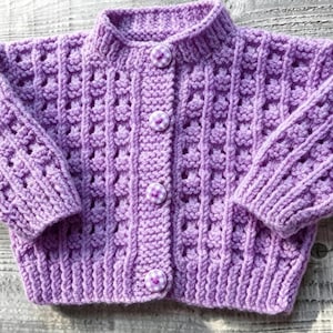 May include: A light purple long-sleeve cardigan featuring a textured waffle-knit pattern, a ribbed collar, and four round buttons with a white and purple checkered fabric covering. The garment is constructed with a thick yarn and has ribbed cuffs.