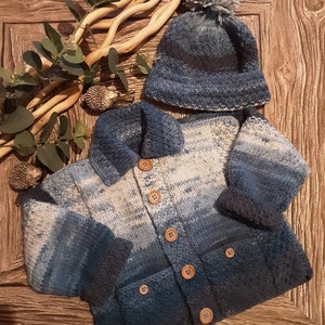 May include: A blue and white knitted cardigan with a button closure and two pockets. The cardigan has a textured knit and a pom-pom on the top of the hat.