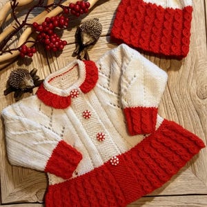 Könnte beinhalten: Ein weiß-roter Strickpullover mit Rundhalsausschnitt, langen Ärmeln und ausgestelltem Saum, mit Zopfstrickmustern und vier runden weißen Knöpfen mit roten Punkten. Eine passende weiß-rote Strickmütze ist ebenfalls enthalten.