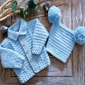 Könnte beinhalten: Eine hellblaue Strickjacke mit Kragen und fünf kleinen Zierknöpfen auf der Vorderseite, kombiniert mit einer passenden hellblauen Strickmütze mit zwei großen Bommeln an den oberen Ecken, wobei beide Teile ein strukturiertes Muster aufweisen.