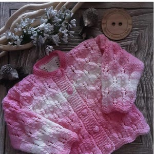 May include: A pink and white knitted cardigan with a button-up front. The cardigan has a textured pattern and is labeled "Oona" with a size range of 18"-22". The garment is displayed on a wooden surface.