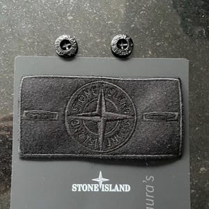 Könnte beinhalten: Ein schwarzer, rechteckiger Stone Island-Aufnäher mit Kompass-Logo, begleitet von zwei schwarzen, runden Knöpfen mit eingeprägtem Stone Island-Markennamen, alles auf einer dunkelgrauen Marken-Präsentationskarte befestigt.